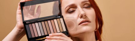 redhead woman holding eye shadow palette near face on beige background, makeup advertisement banner