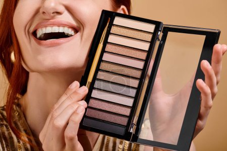 cropped view of happy woman holding eye shadow palette on beige background, makeup advertisement