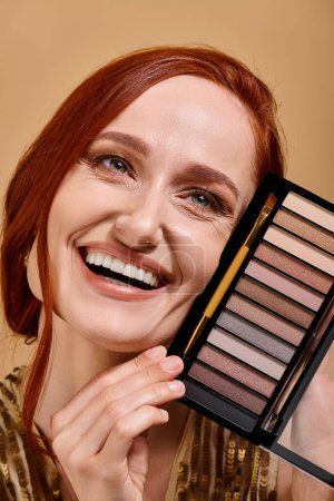 primer plano de la mujer feliz sosteniendo la paleta de sombra de ojos sobre fondo beige, publicidad de maquillaje