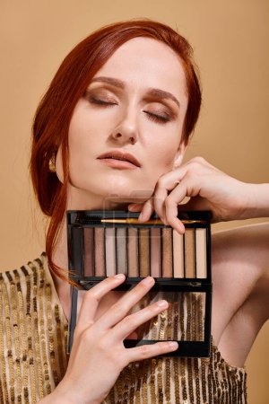 redhead woman holding eye shadow palette near face on beige background, beauty advertisement