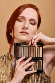redhead woman holding eye shadow palette near face on beige background, beauty advertisement Poster #693713846