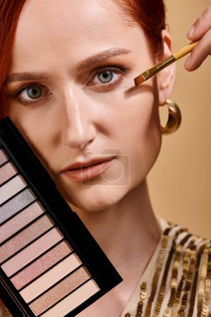 close up of redhead woman applying eye shadow and holding palette on beige backdrop, pastel color
