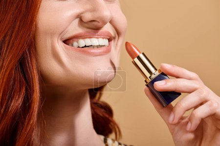 vista recortada de mujer pelirroja sonriendo y aplicando lápiz labial desnudo sobre fondo beige, producto de maquillaje