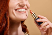 cropped view of redhead woman smiling and applying nude lipstick on beige background, makeup product t-shirt #693713860