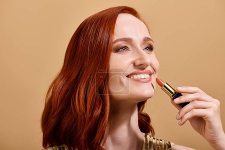 Fröhliche und rothaarige Frau lächelt und trägt Nude-Lippenstift auf beigem Hintergrund auf, Make-up-Produkt