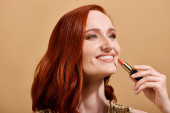 Joyful and redhead woman smiling and applying nude lipstick on beige background, makeup product mug #693713866