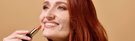 Joyful and redhead woman smiling and applying nude lipstick on beige background, makeup banner