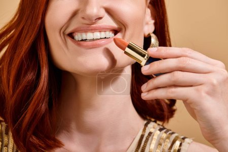 partial view of woman smiling and applying nude lipstick on beige background, makeup product