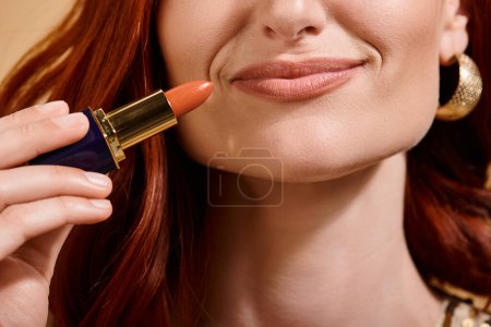 cropped view of woman smiling and applying nude lipstick on beige background, makeup product