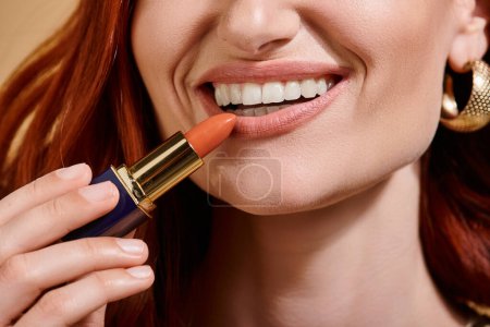vista recortada de la mujer sonriendo y aplicando lápiz labial desnudo sobre fondo beige, dientes blancos
