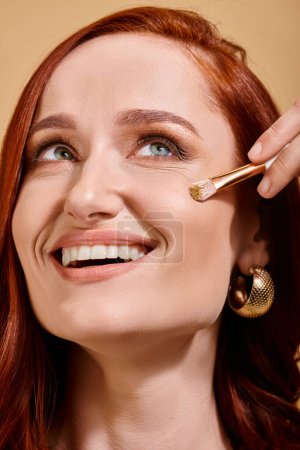 Radiant and redhead woman applying sparkling glitter on cheek with makeup brush on beige backdrop