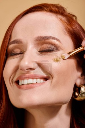 Mujer radiante y pelirroja aplicando brillo brillante en la mejilla con cepillo de maquillaje sobre fondo beige