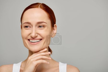 cheerful and redhead woman in her 30s posing in white tank top on grey background, genuine smile