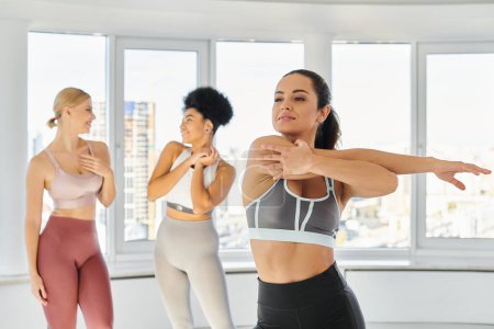 brunette woman in sportswear stretching before pilates class next to interracial female friends
