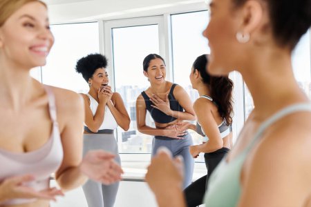 focus on diverse female friends in sportswear laughing while chatting before pilates class