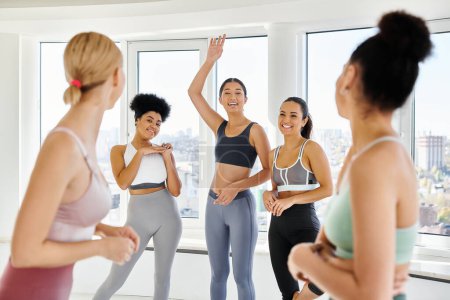 focus on asian woman in sportswear waving at her female friends in pilates class, diverse group