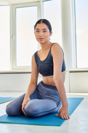 junge und brünette asiatische Frau in ihren Zwanzigern, die auf einer Yogamatte sitzt und in die Kamera schaut, sportlich