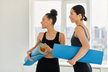 Happy and young women in their 20s standing with yoga mats and smiling in studio, post-workout