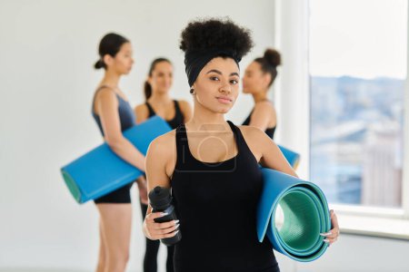 focus on african american woman standing with fitness mat and sports bottle in yoga studio