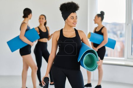 Foto de Enfoque en la mujer afroamericana feliz de pie con tapete de fitness y botella de deportes en el estudio de yoga - Imagen libre de derechos