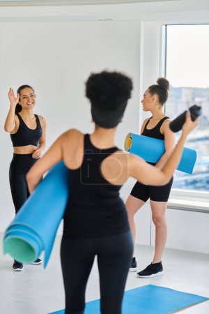 happy brunette woman waving hands at her african american friend with fitness mat in yoga studio