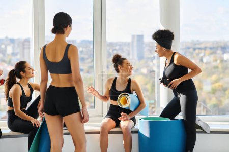 happy multicultural young women in 20s chatting while sitting next to window in pilates studio
