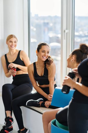 Téléchargez les photos : Joyeuses jeunes femmes interraciales dans la vingtaine bavardant tout en étant assis à côté de la fenêtre dans un studio de yoga - en image libre de droit