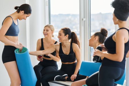 cheerful interracial young women in 20s chatting while sitting next to window in pilates studio