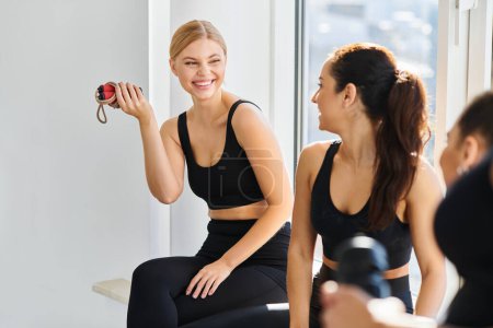 glückliche blonde Frau mit Springseil lacht, während sie mit ihrem Freund im Pilates-Studio plaudert