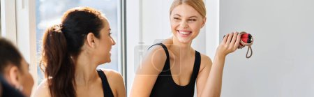 happy blonde woman with jumping rope laughing while chatting with friend in pilates studio, banner