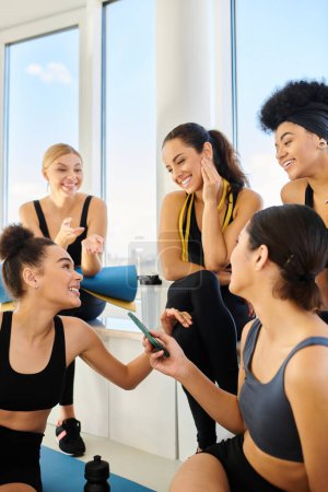 group of five multicultural woman in sportswear chatting after workout in pilates studio