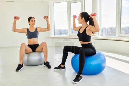 happy diverse female friends exercising with dumbbells on fitness balls during pilates class
