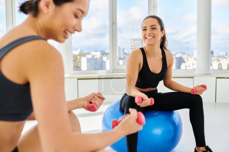 happy interracial female friends exercising with dumbbells on fitness balls during pilates class