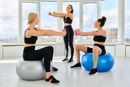 joyful multicultural female friends exercising with dumbbells on fitness balls near trainer
