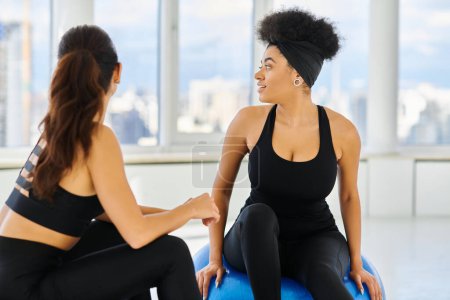 happy and diverse female friends sitting on fitness balls and looking at window during pilates class