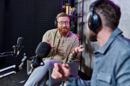 bärtiger gutaussehender Interviewer und sein Gast mit Kopfhörern im Studio diskutieren Fragen, Podcast