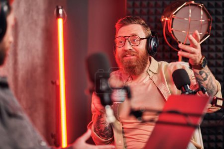 fröhlicher gutaussehender Mann mit rotem Bart und Kopfhörer in lässiger Kleidung beim Podcast-Gespräch