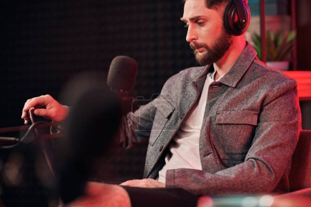 bel homme barbu avec écouteurs en tenue décontractée assis pendant podcast en studio