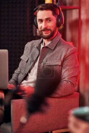 hombre barbudo guapo con auriculares en traje casual sentado durante el podcast, mientras que en el estudio