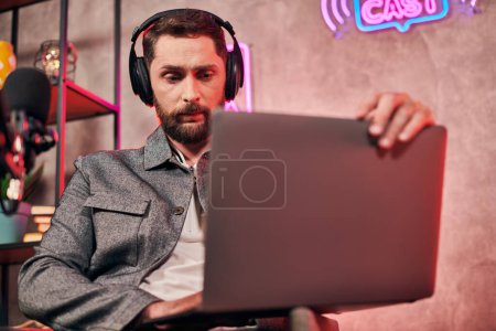 gut aussehender bärtiger Mann in bequemer Alltagskleidung sitzt mit Laptop beim Podcast im Studio