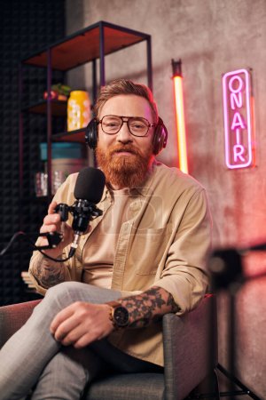 alegre hombre guapo con el pelo rojo y la barba con auriculares mirando a la cámara durante el podcast
