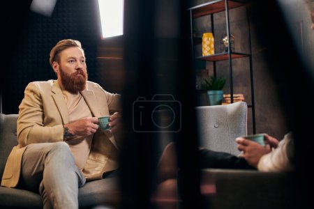 Photo for Handsome bearded man with red hair in elegant clothes sitting next to his interviewer in studio - Royalty Free Image