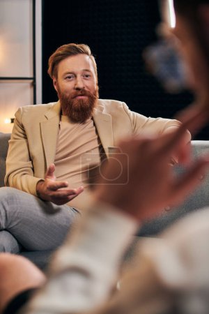 Photo for Good looking bearded man with red hair in elegant clothes sitting next to his interviewer in studio - Royalty Free Image