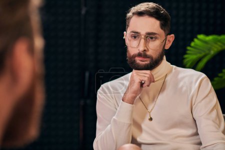 handsome bearded man with glasses in elegant outfit sitting and looking at his interviewer