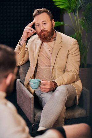 good looking bearded man with coffee cup sitting during discussion next to his interviewer