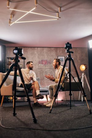 dos hombres elegantes con barbas con estilo elegante sentado y discutiendo preguntas de la entrevista en el estudio
