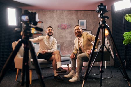hombres alegres de buen aspecto en trajes elegantes sonriendo felizmente a la cámara durante la entrevista en el estudio