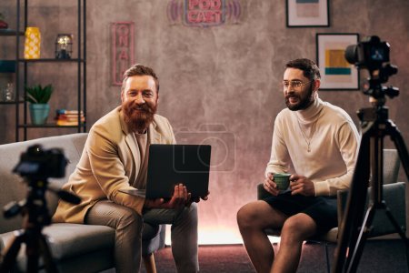 Foto de Hombres guapos alegres en trajes de moda con café y portátil sonriendo durante la entrevista - Imagen libre de derechos