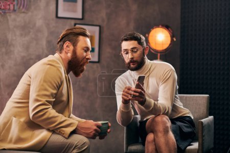 two handsome elegant men with beards in chic clothes looking at smartphone during interview