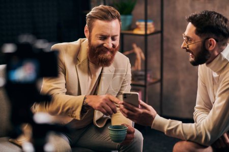 two cheerful elegant men with beards in chic clothes looking at smartphone during interview